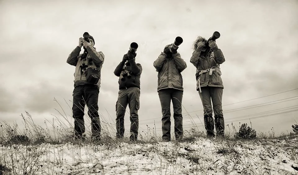 Cztery osoby stoją na lekko ośnieżonym terenie trzymając lornetki lub aparaty z teleobiektywami i patrząc w dal, pochmurne niebo w tle.