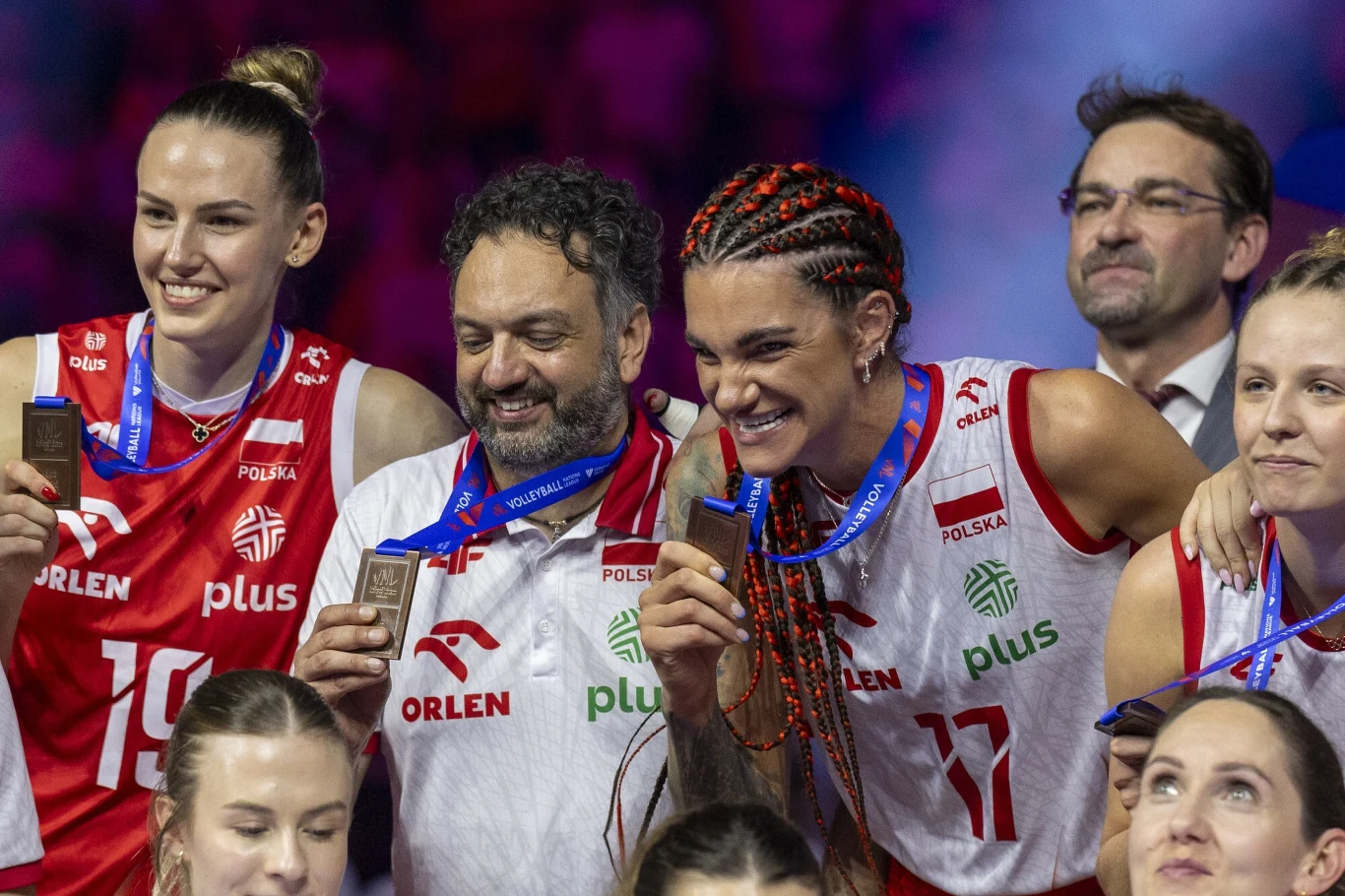 Zawodniczki reprezentacji Polski w siatkówce oraz członkowie sztabu trzymają medale i świętują sukces podczas sportowej uroczystości, uśmiechając się do kamery.