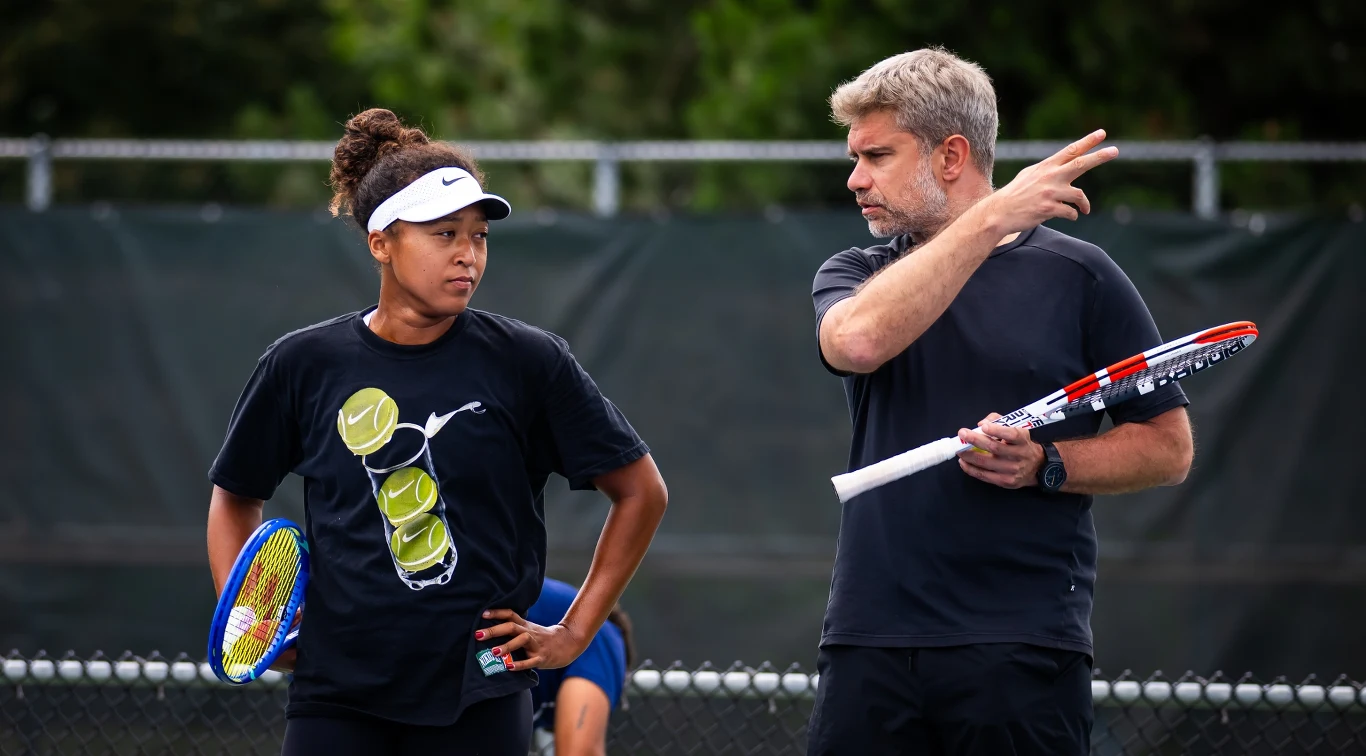Naomi Osaka i Tomasz Wiktorowski Dwie osoby stoją na korcie tenisowym, jedna z nich trzyma rakietę tenisową i jest ubrana w czarną koszulkę z nadrukiem piłek tenisowych, druga osoba w czarnym stroju sportowym trzyma rakietę i gestykuluje, jakby tłumacząc strategię gry.