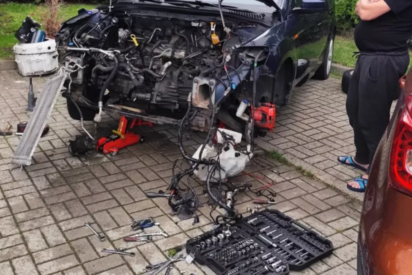 Strażników miejskich o naprawie na parkingu i wyciekającym płynie poinformował jeden z mieszkańców. Samochód z otwartą maską oraz wymontowanym silnikiem, rozłożonymi częściami i licznymi narzędziami warsztatowymi na ziemi, obok stoi mężczyzna w czarnej odzieży.