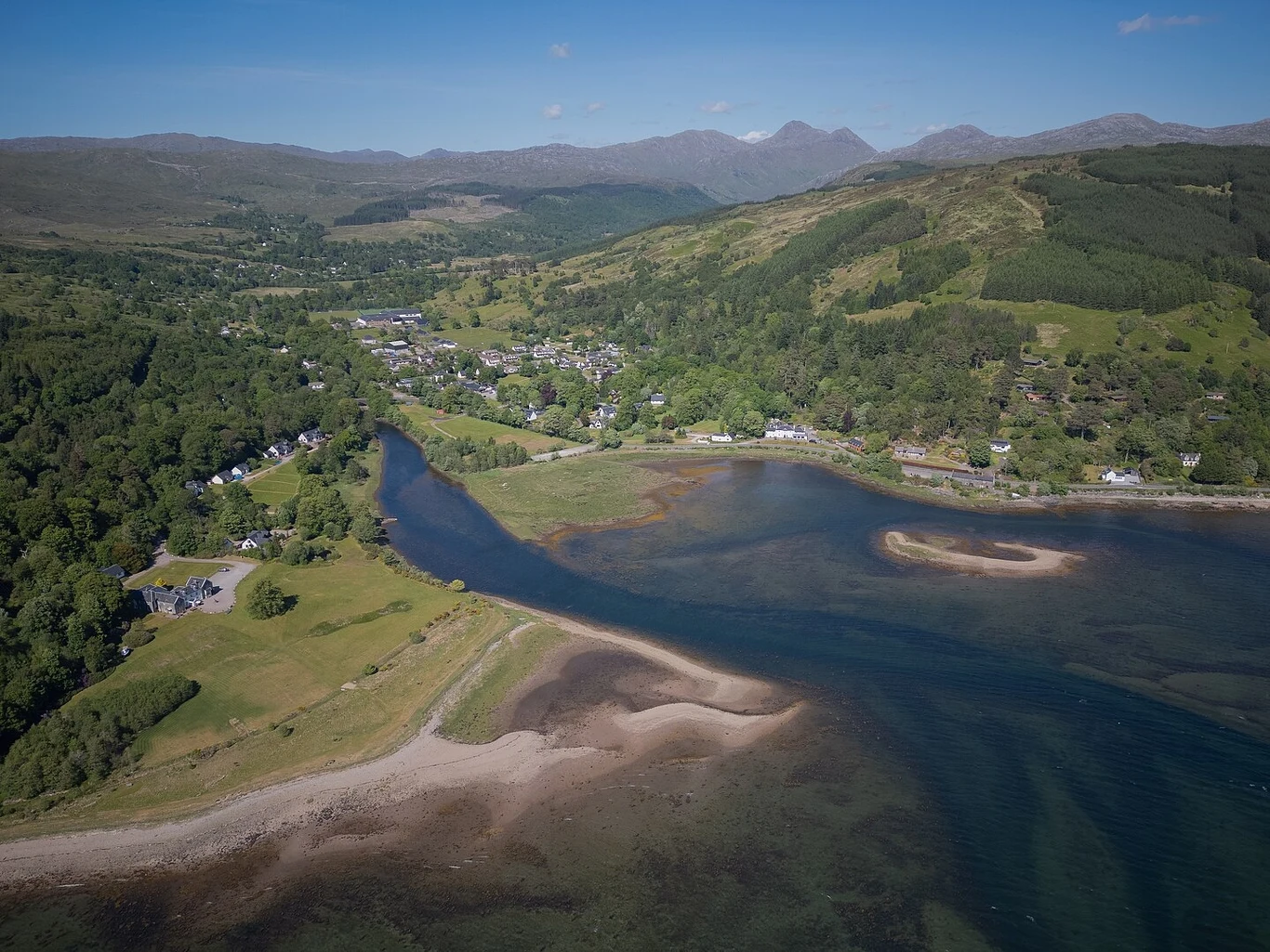 Niedaleko wioski Strontian na wodach Loch Sunart znajduje się malownicza wyspa Eilean Mòr. Wystawiono ją na sprzedaż. Widok na zielone wzgórza oraz niewielką zatokę z łagodnie opadającą linią brzegową, w tle zabudowania wśród drzew oraz góry na horyzoncie.