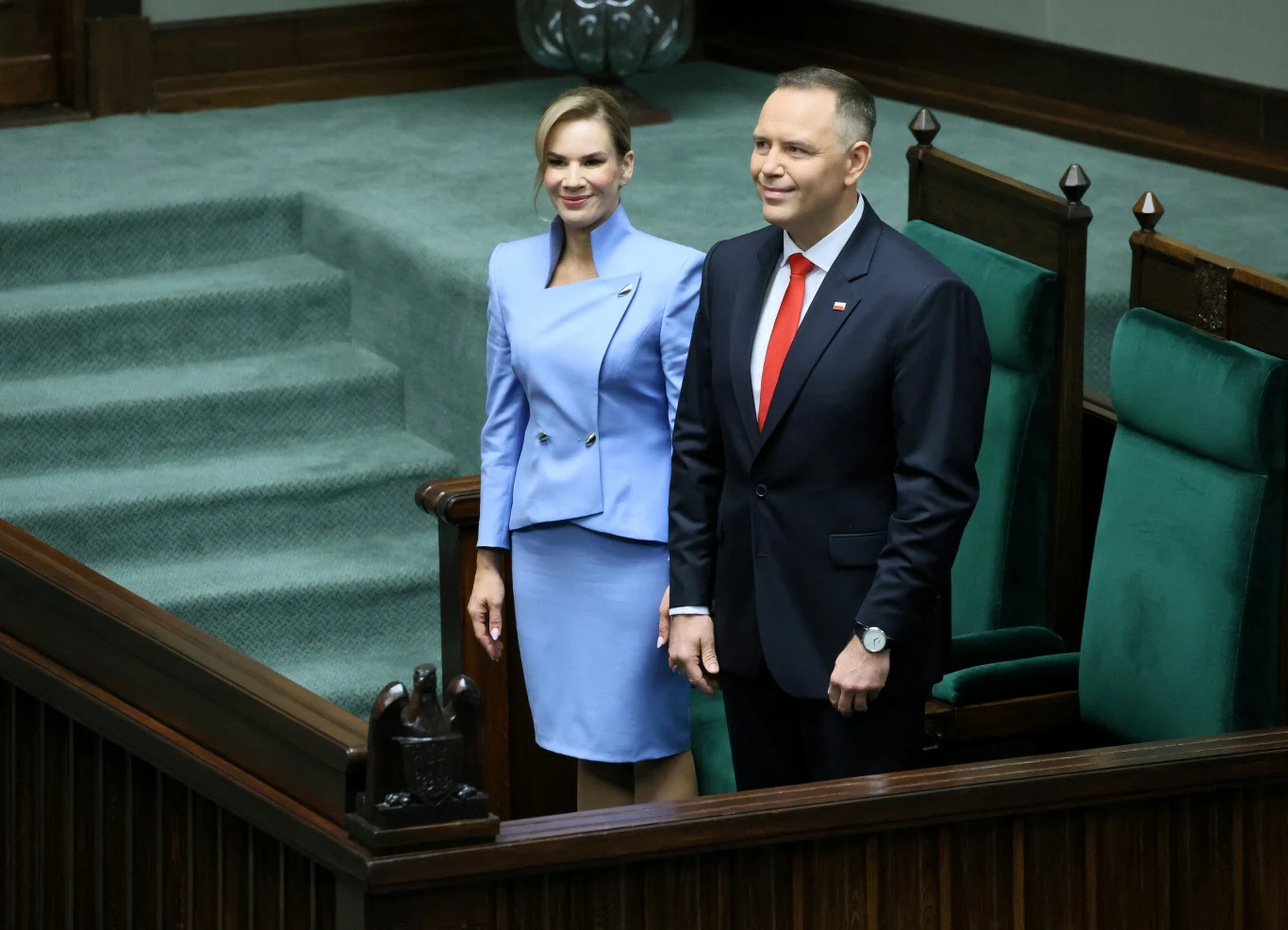 Para w eleganckich, formalnych strojach stoi obok siebie w sali sejmowej, przy charakterystycznych, zielonych ławach parlamentarnych, tło stanowią schody wykładane wykładziną i ozdobne elementy architektoniczne.