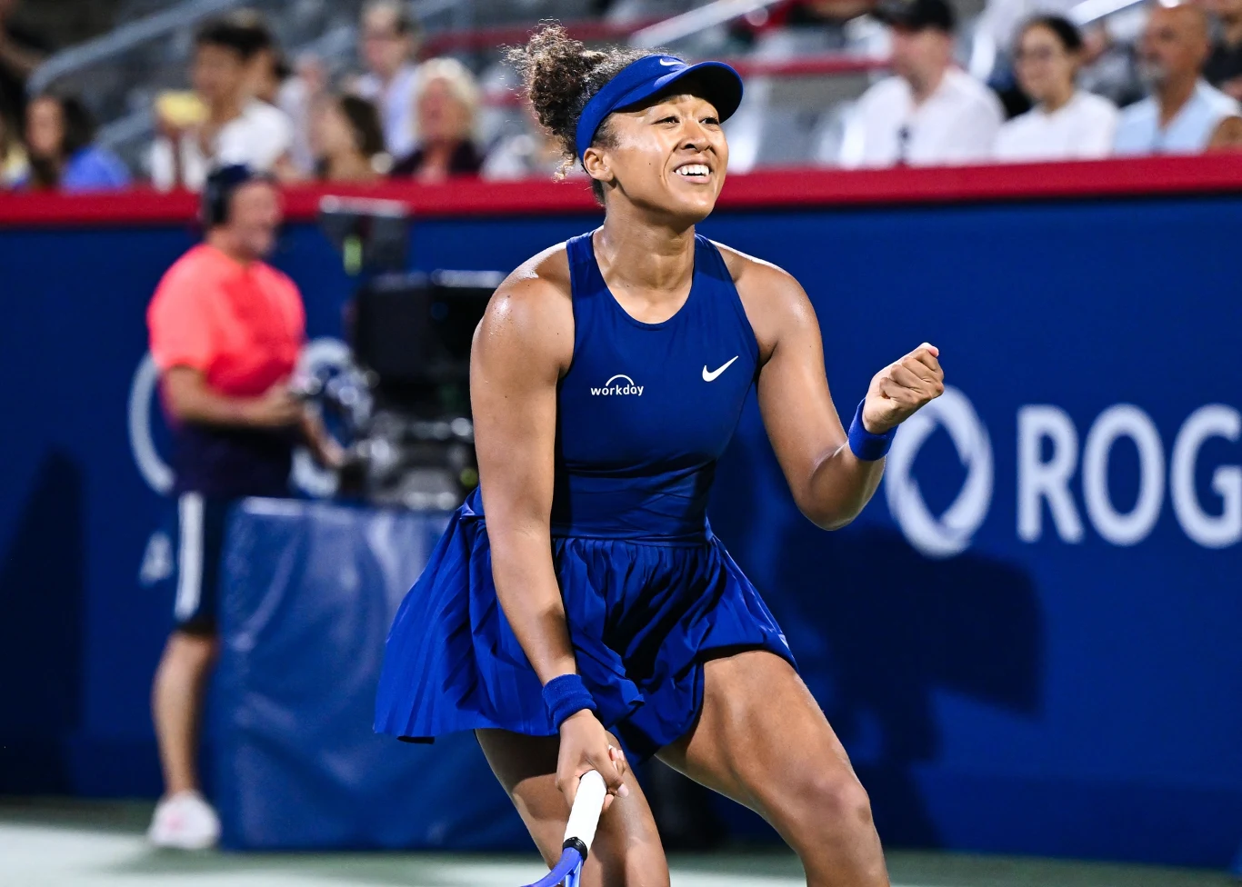 Naomi Osaka Tenisistka w niebieskim stroju sportowym świętuje zdobycie punktu na korcie, w tle widownia i elementy oznaczenia turnieju.