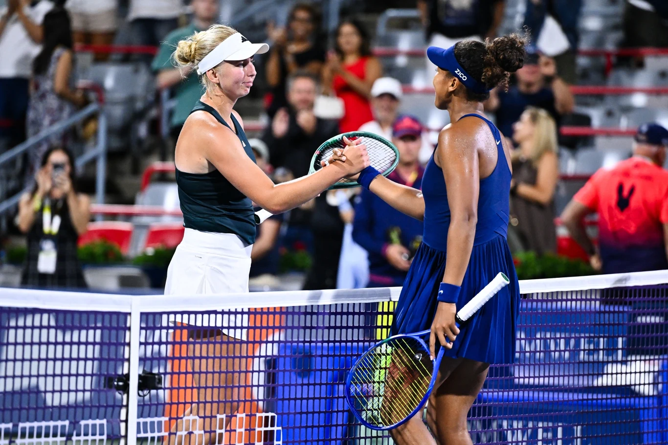 Clara Tauson i Naomi Osaka Dwie tenisistki podają sobie ręce przez siatkę na korcie, obie trzymają rakiety, wokół widoczna publiczność i sędziowie na tle trybun.