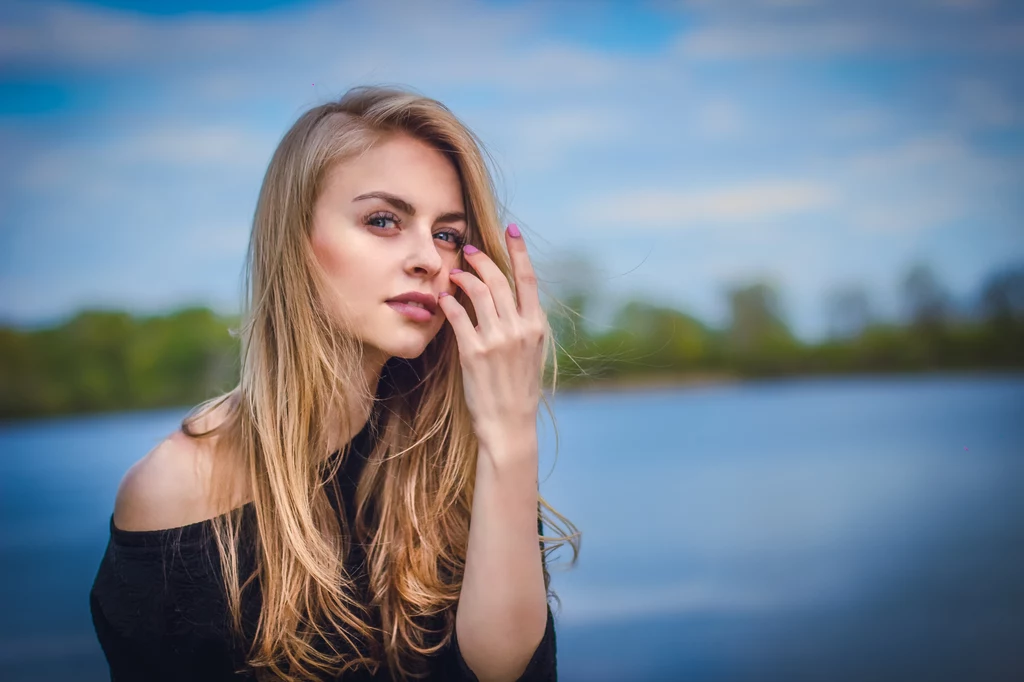 Młoda kobieta o długich blond włosach patrzy prosto w obiektyw, przytrzymując włosy dłonią, w tle rozmyte jezioro i drzewa oraz błękitne niebo z chmurami.