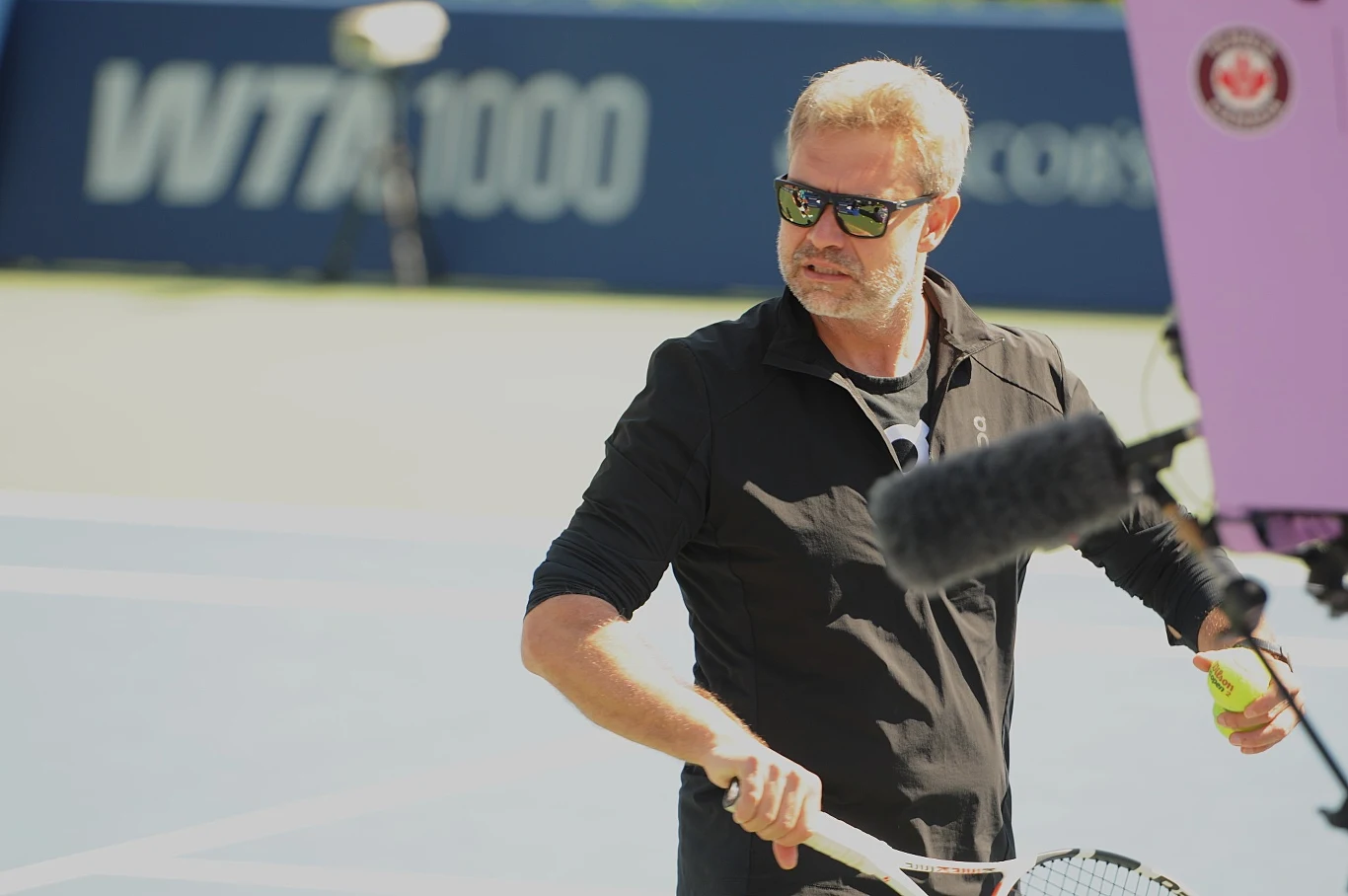 Tomasz Wiktorowski Mężczyzna w okularach przeciwsłonecznych trzymający rakietę tenisową oraz piłkę, stojący na korcie tenisowym; na pierwszym planie widoczny mikrofon, w tle niebieska mata z napisem WTA1000.