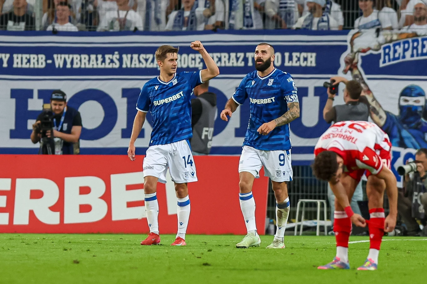 Leo Bengtsson i Mikael Ishak cieszący się z gola Dwóch zawodników w niebieskich koszulkach świętuje zdobycie gola podczas meczu piłkarskiego, obok gracz drużyny przeciwnej pochyla się w geście rozczarowania; w tle widoczni kibice z transparentami.