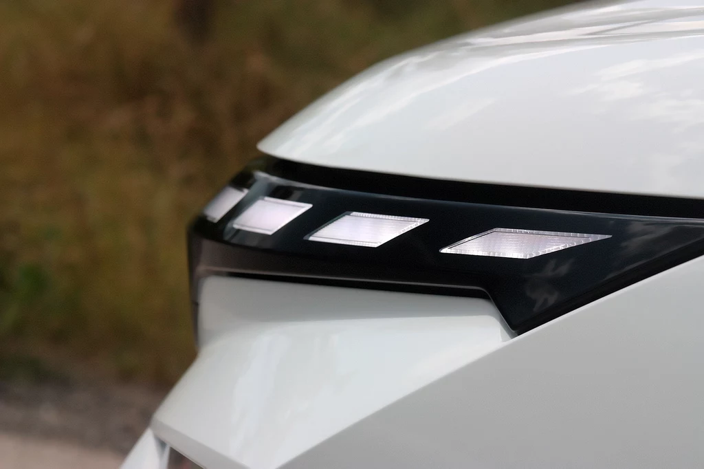 Światła do jazdy dziennej są całkowicie nowe Przedni reflektor nowoczesnego, białego samochodu z wyraźnym, czarnym elementem dekoracyjnym i zintegrowanymi światłami LED, na tle rozmytego, zielonego otoczenia.
