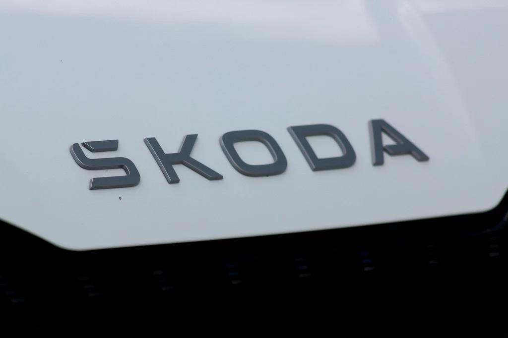 Na masce Enyaq po liftingu nie znajdziemy już loga Skody, który został zastąpiony napisem Logo marki Škoda na karoserii białego samochodu, wyraźnie widoczne litery na tle jasnej powierzchni.