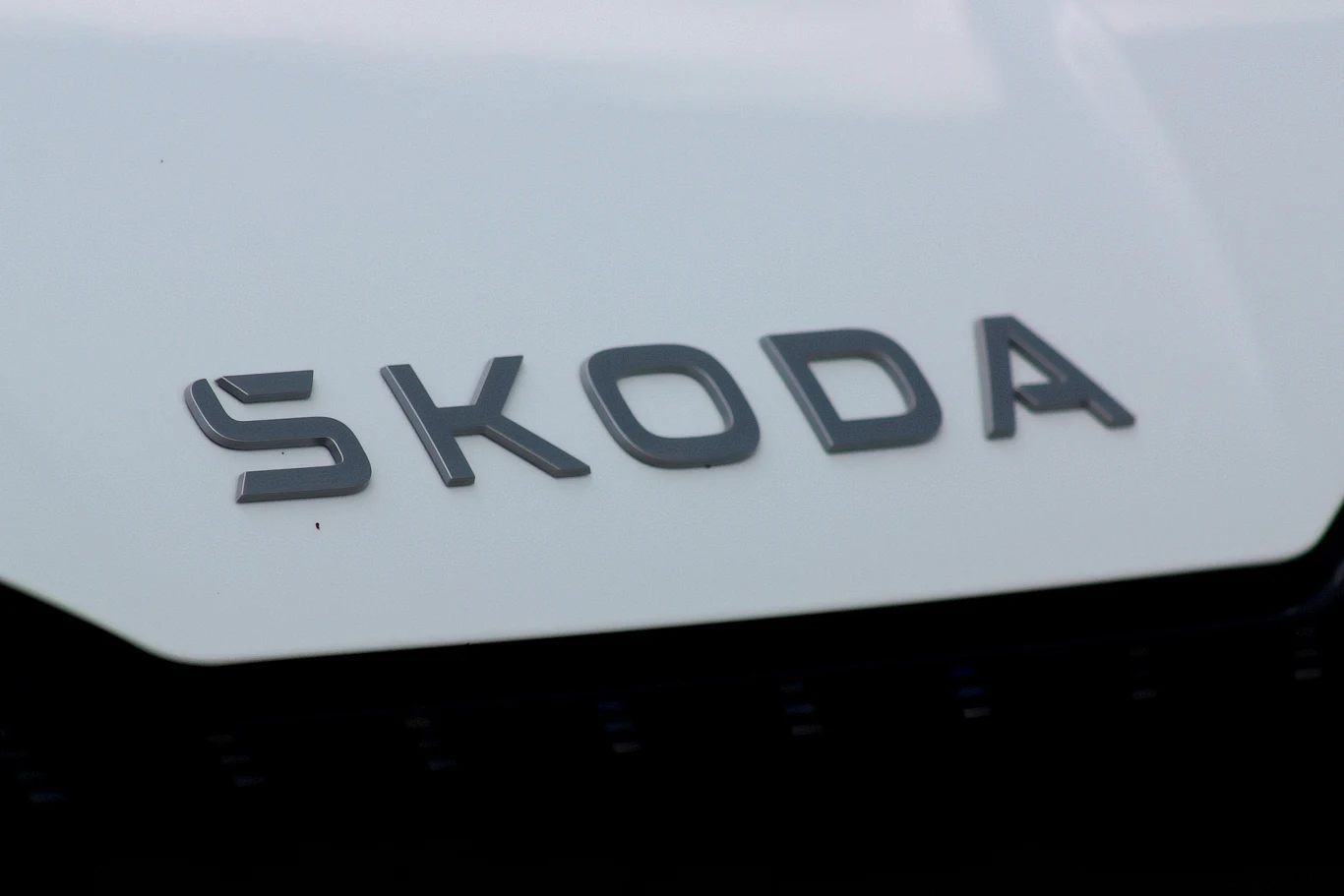 Logo marki Škoda na karoserii białego samochodu, wyraźnie widoczne litery na tle jasnej powierzchni.