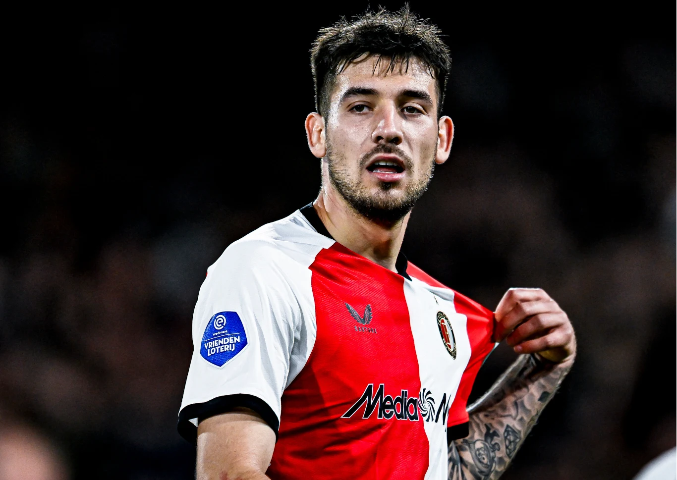 Jakub Moder Piłkarz w czerwono-białej koszulce, trzymający za kołnierz swojego stroju, z widocznym logo sponsora oraz emblematem ligi na rękawie, na ciemnym, rozmytym tle stadionu.