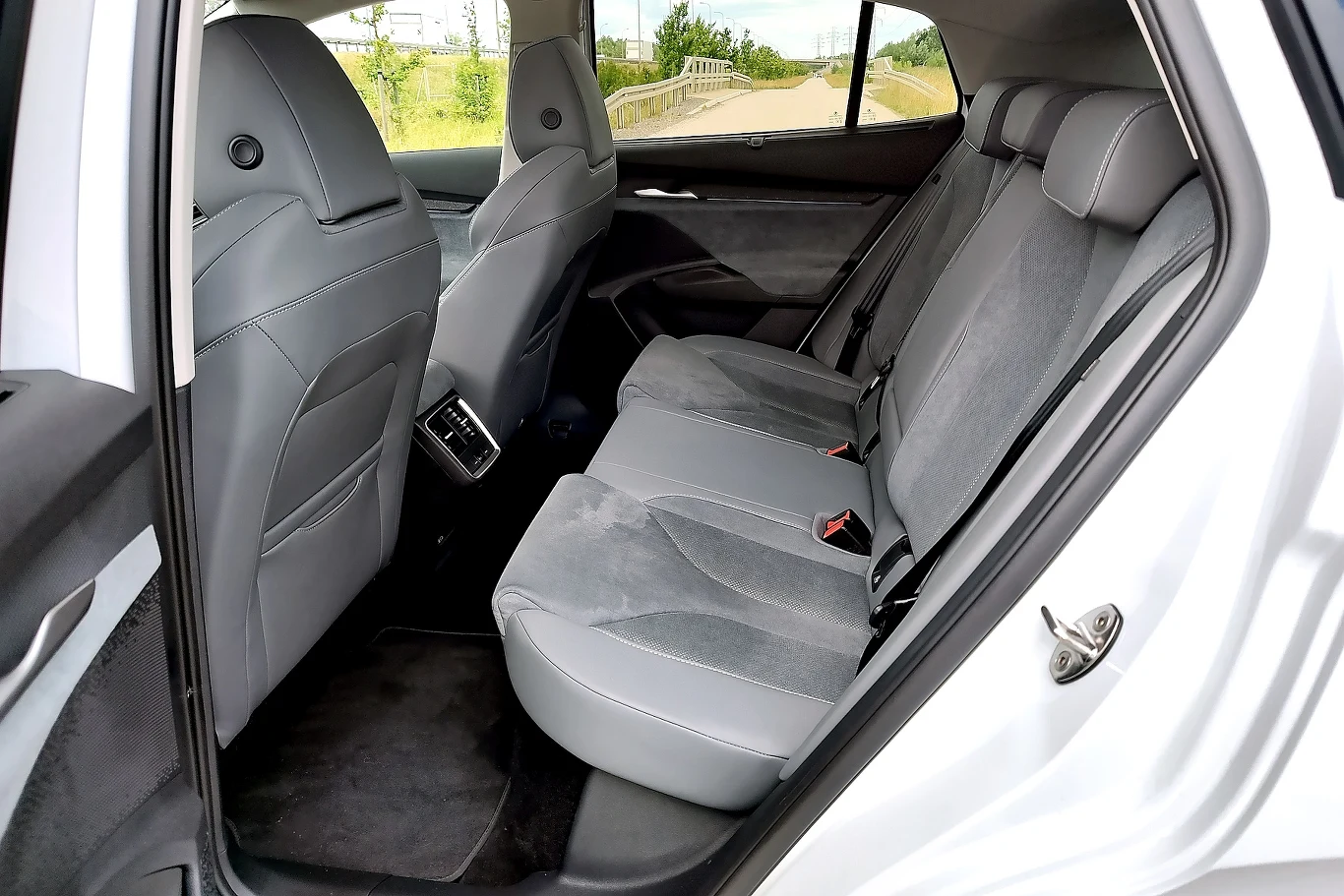 Widok wnętrza samochodu z tyłu, trzy szare tapicerowane fotele tylnej kanapy, jasna wyściółka i elementy drzwi, przestronne miejsce na nogi, fragmenty przednich siedzeń oraz otwarte drzwi po lewej stronie, przez okna widoczna zieleń.