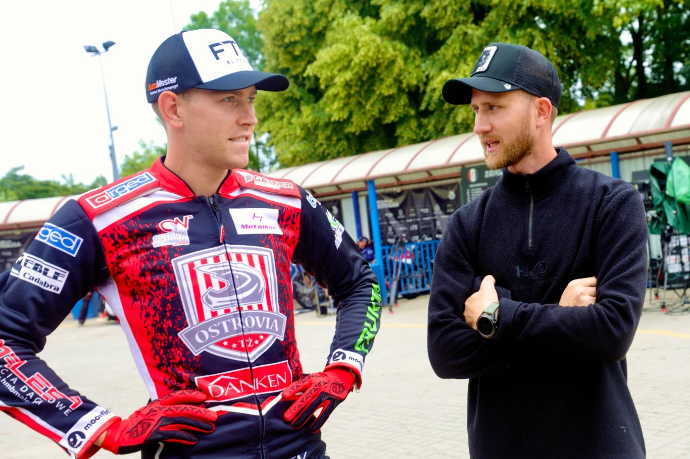 Fredrik Jakobsen w rozmowie z Mikkelem Bechem Dwóch mężczyzn rozmawia na zewnątrz, jeden ubrany w kombinezon sportowy z logo Ostrovia, drugi w czarną kurtkę i czapkę, w tle widoczne zabudowania oraz drzewa.