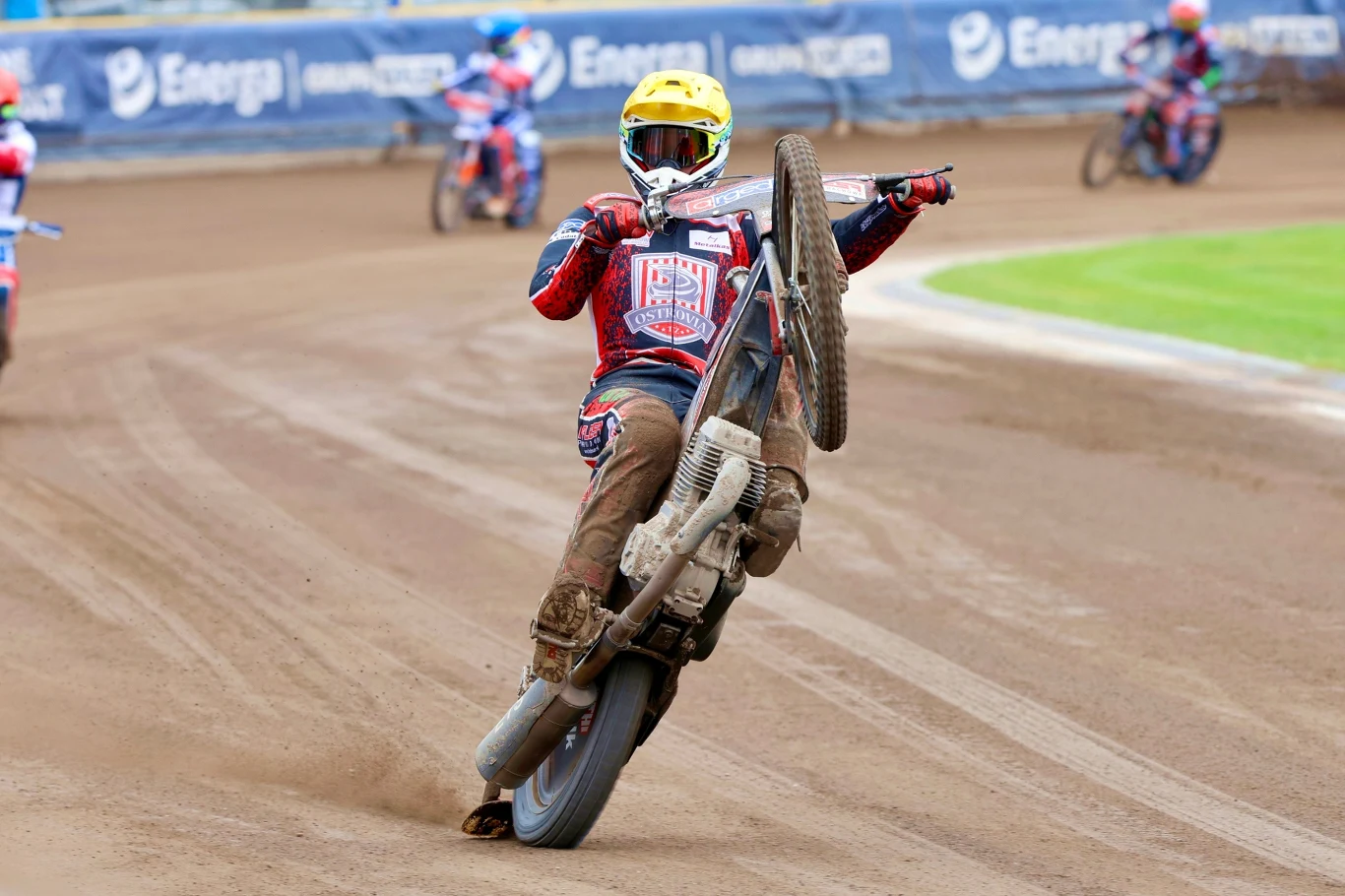 Fredrik Jakobsen Zawodnik sportów motorowych w kolorowym kombinezonie i żółtym kasku wykonuje efektowny manewr jazdy na jednym kole motocyklem żużlowym na torze, w tle widoczni są inni uczestnicy wyścigu oraz fragmenty reklamy na ogrodzeniu stadionu.