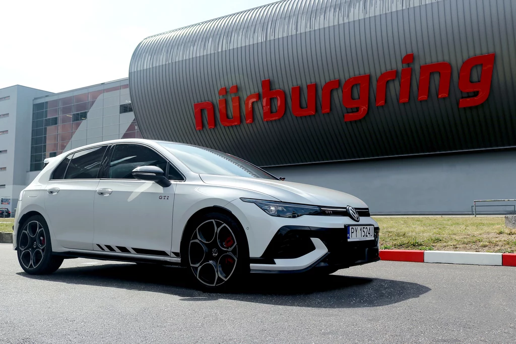 Tor Nürburgring Nordschleife wydaje się naturalnym wyborem do przetestowania Golfa GTI Clubsport. Biały sportowy samochód Volkswagen Golf GTI stoi zaparkowany przed charakterystycznym budynkiem toru wyścigowego Nürburgring z dużym czerwonym napisem na tle metalowej konstrukcji.
