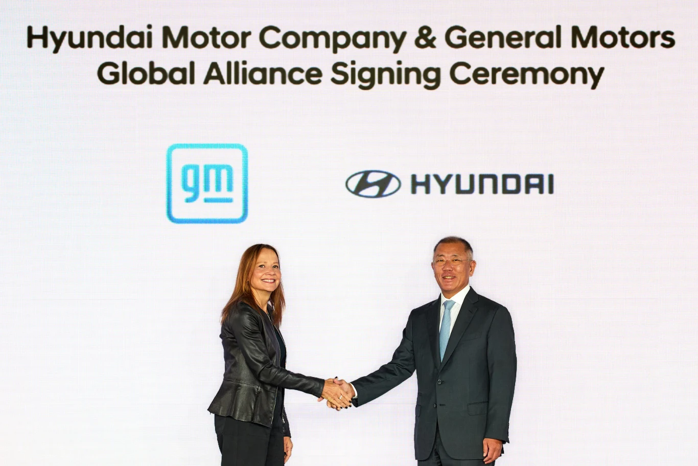 Dwie osoby w garniturach wymieniają uścisk dłoni na tle białej ściany z logotypami General Motors oraz Hyundai i napisem informującym o ceremonii podpisania globalnego sojuszu.