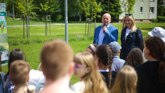 Mężczyzna w garniturze trzymający mikrofon oraz kobieta stoją razem przed grupą dzieci na świeżym powietrzu, w tle tablica edukacyjna i zieleń miejska.