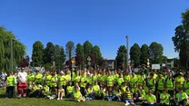 Duża grupa dzieci i dorosłych ubrana w żółte kamizelki odblaskowe stoi na trawniku, w tle drzewa oraz zabudowania miejskie, nad głowami czyste niebo.