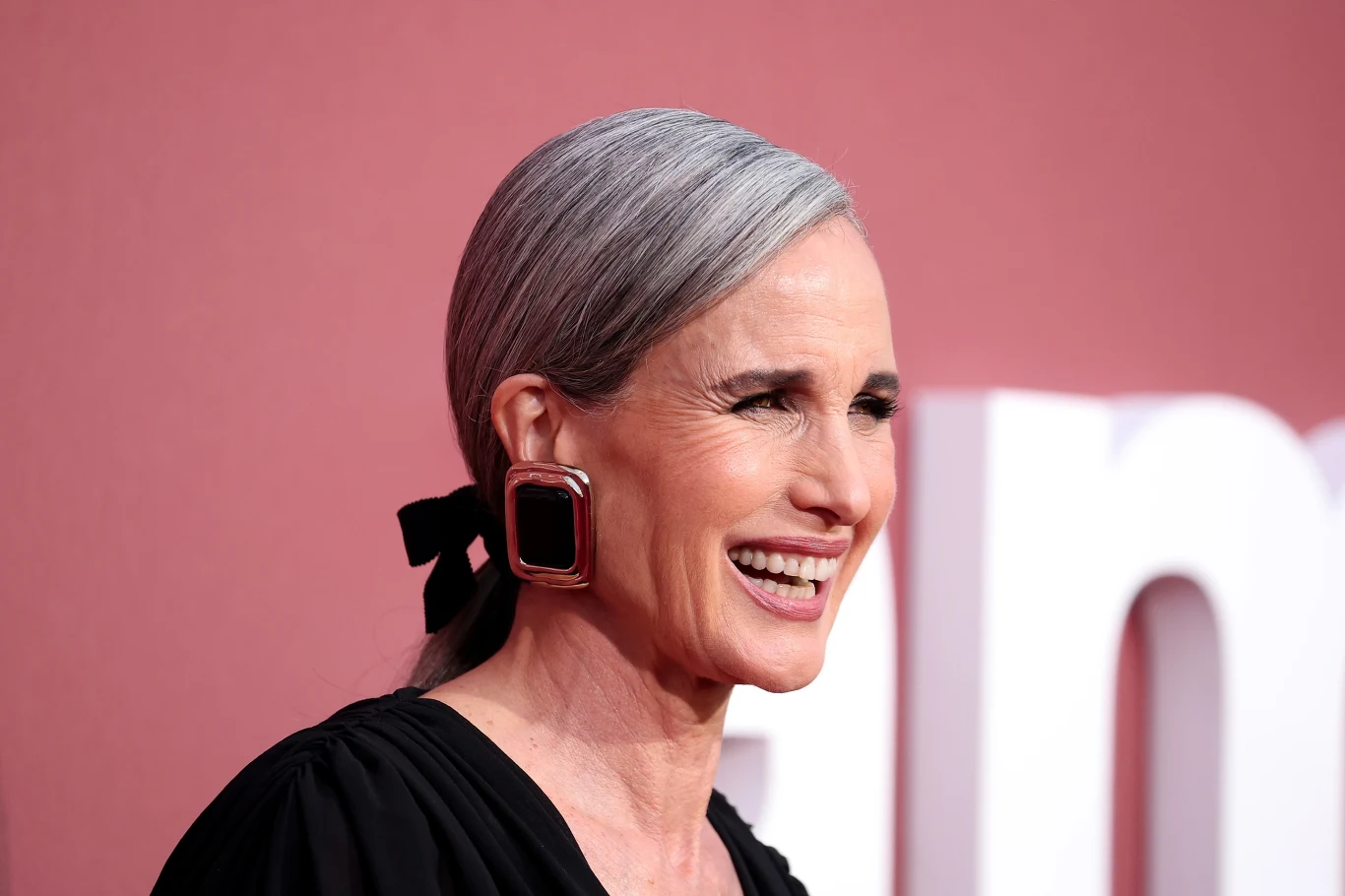 Andie MacDowell z dumą eksponuje siwe włosy Uśmiechnięta kobieta z siwymi włosami spiętymi do tyłu, w dużych kwadratowych kolczykach i czarnej bluzce, na tle różowej ściany z białym elementem napisu.