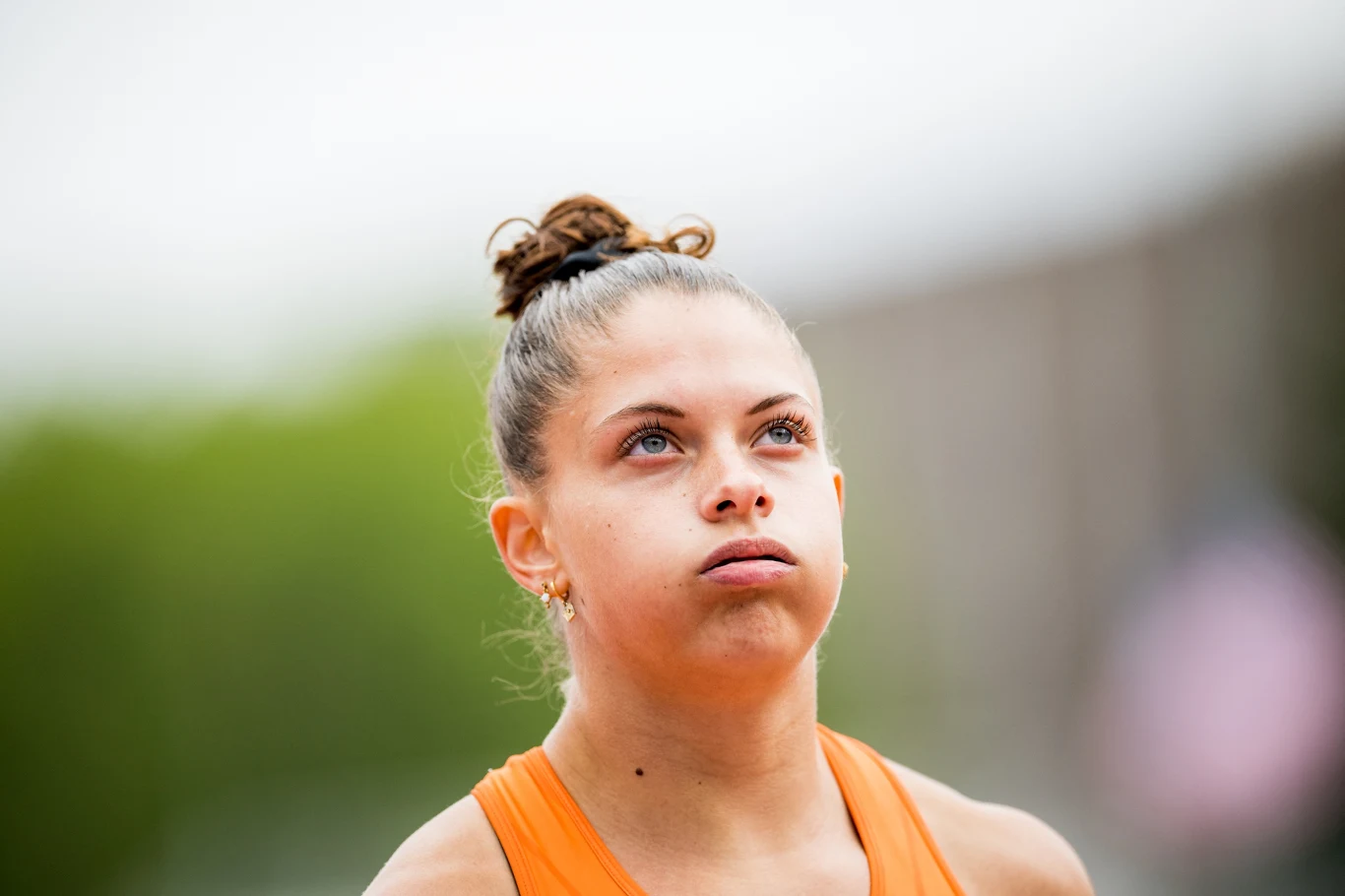 Młoda kobieta w sportowej koszulce pomarańczowej z zaplecionymi włosami patrzy do góry z poważnym wyrazem twarzy, na rozmytym tle na zewnątrz.
