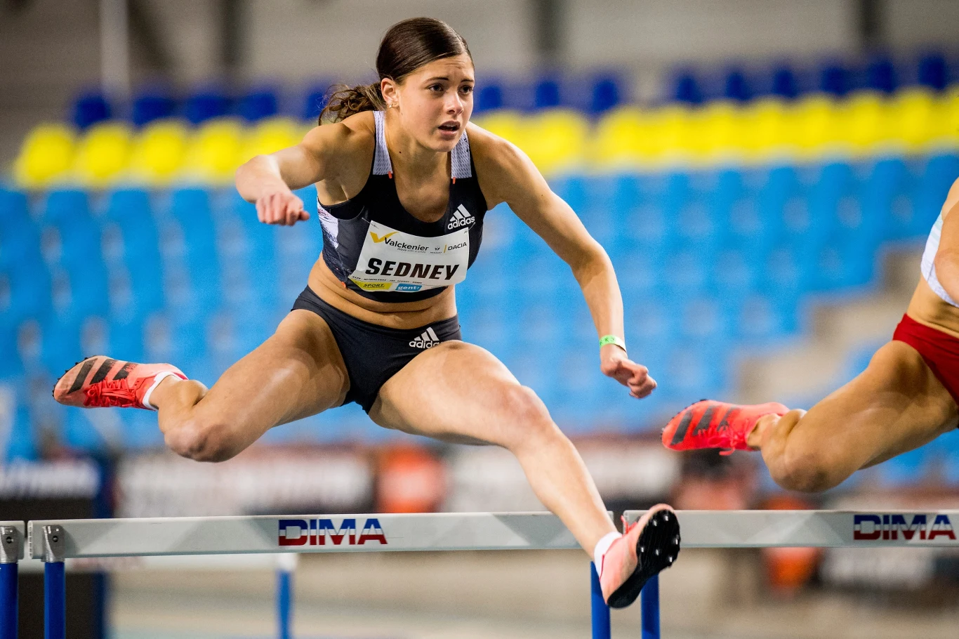 Zawodniczka lekkoatletyczna podczas biegu przez płotki w hali sportowej, dynamiczny skok nad przeszkodą, widoczne puste trybuny w tle, intensywna koncentracja na twarzy sportowczyni.