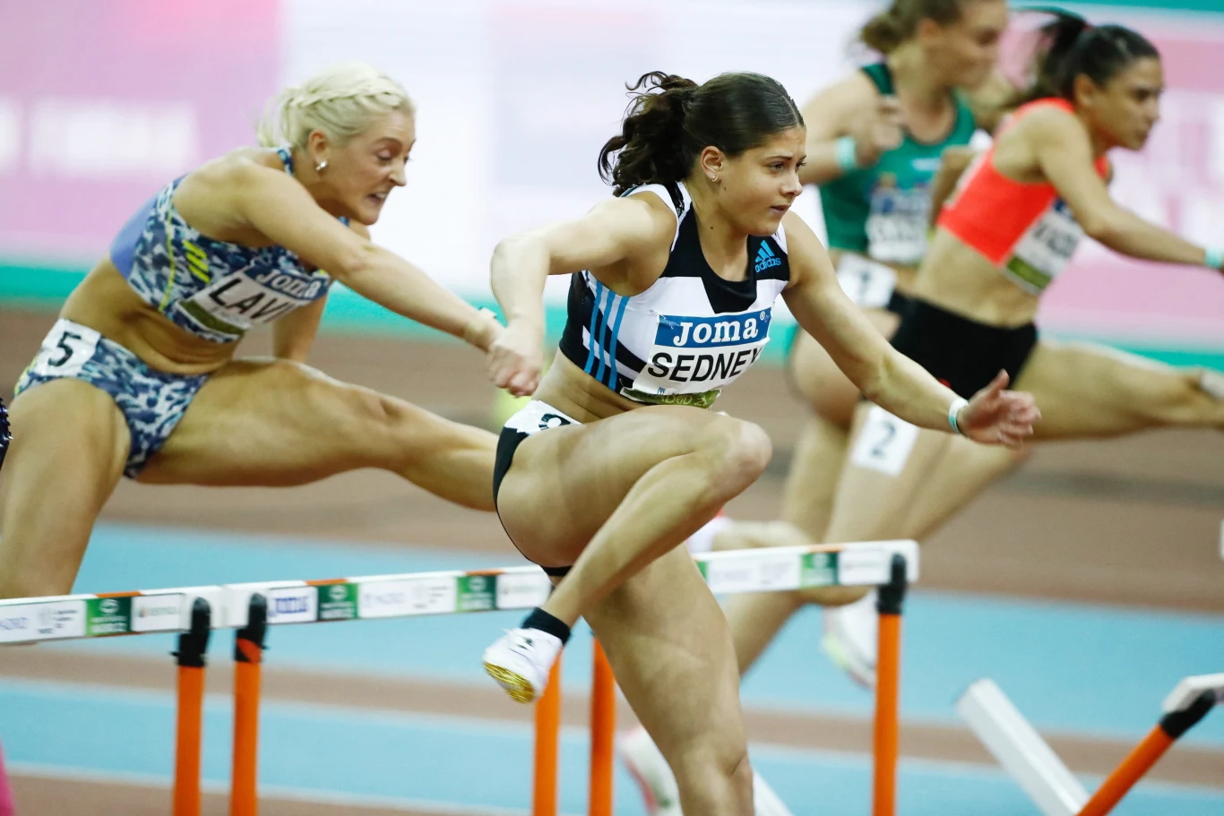Zawodniczki pokonujące kolejne płotki podczas wyścigu lekkoatletycznego w hali, dynamiczne ujęcie z rywalizacją na pierwszym planie.