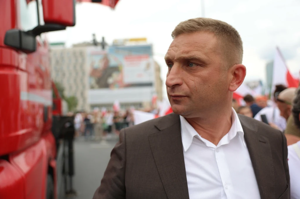 Robert Bąkiewicz dostał wezwanie do prokuratury Mężczyzna w marynarce i białej koszuli stoi na tle czerwonego pojazdu, prawdopodobnie ciężarówki, wśród tłumu z flagami; w oddali widoczne są budynki oraz rozmazane sylwetki innych osób.