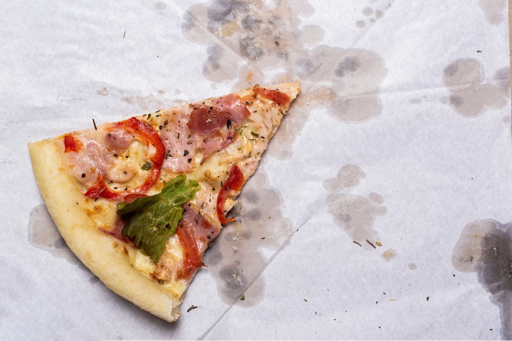 Trójkątny kawałek pizzy z szynką, papryką i liściem sałaty, leżący na papierze z widocznymi tłustymi plamami.
