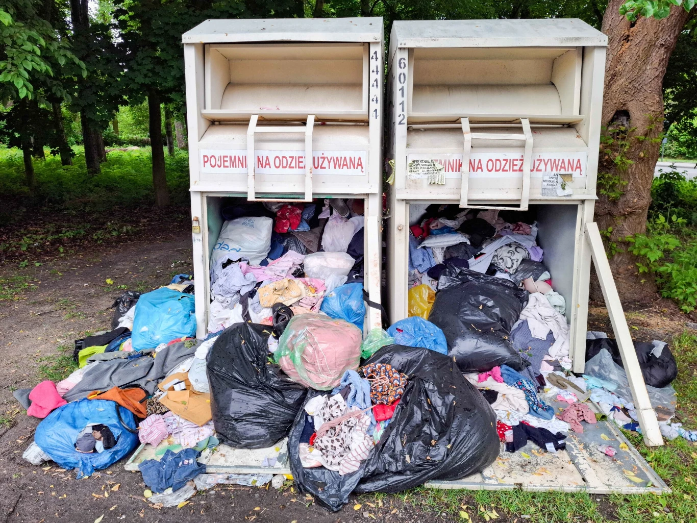 Z powodu nowych przepisów kontenery na ubrania dosłownie pękają w szwach Dwa przepełnione pojemniki na odzież używaną, wokół których rozrzucone są worki i stosy ubrań, otoczone zielenią drzew.
