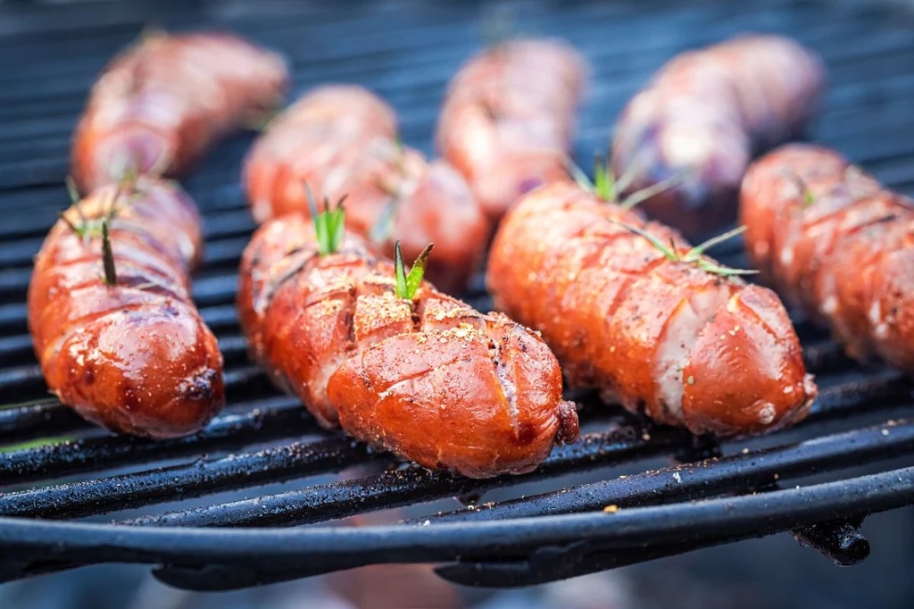 Zbliżenie na dobrze wypieczone kiełbaski grillowane na ruszcie, niektóre z nich udekorowane gałązkami świeżych ziół; na kiełbaskach widać paski przypieczenia i soczystą skórkę.