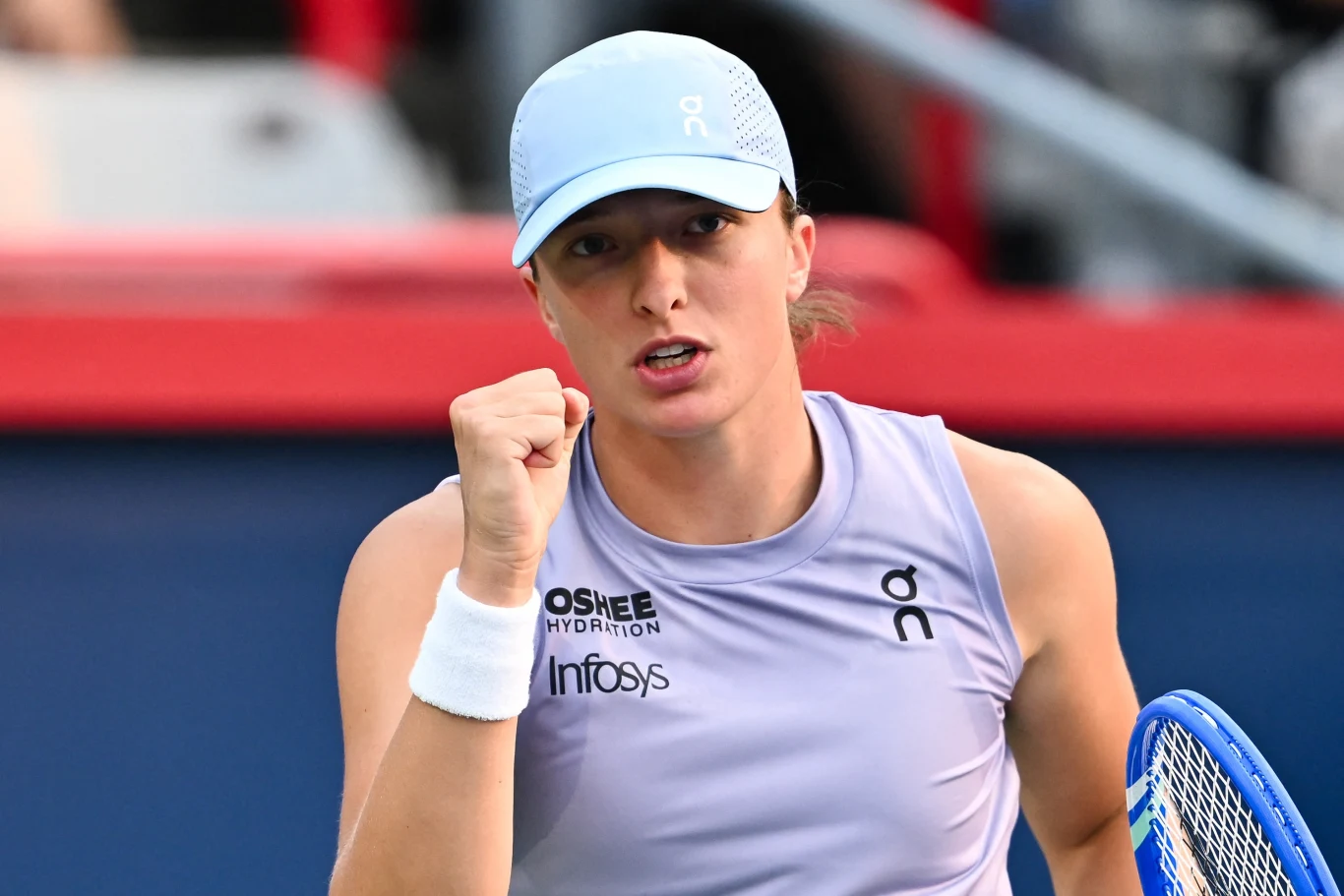 Iga Świątek Tenisistka ubrana w sportowy top i czapkę, ściska pięść w geście triumfu podczas meczu, w tle rozmyte trybuny.