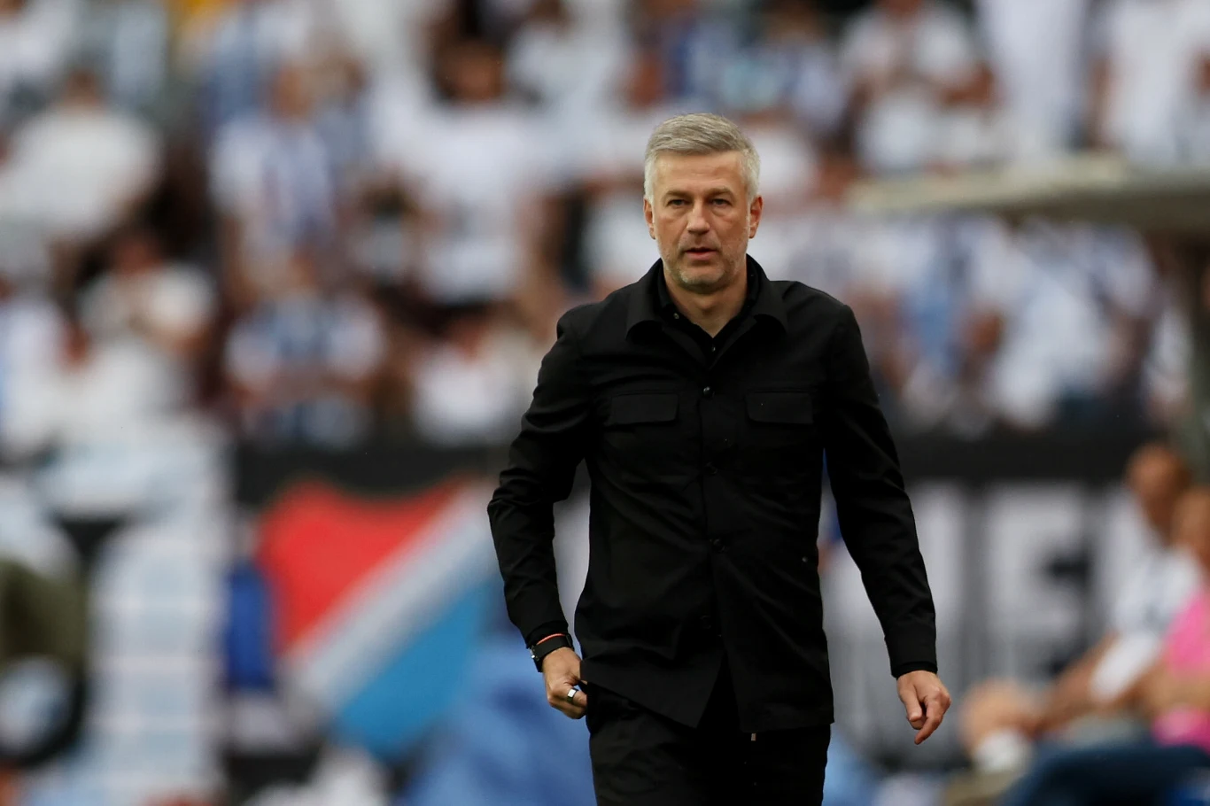 Edward Iordanescu Mężczyzna w czarnym ubraniu stoi na tle stadionu sportowego, wśród rozmytego tłumu widzów.