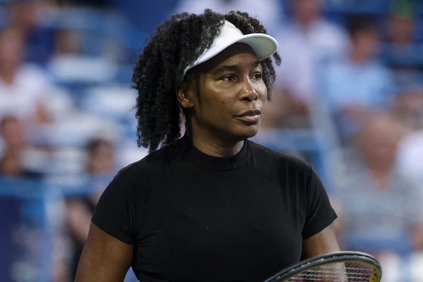 Venus Williams Tenisistka w czarnej koszulce i białej czapce z daszkiem stoi na korcie trzymając rakietę tenisową, skupiona podczas meczu, na tle rozmytej publiczności.