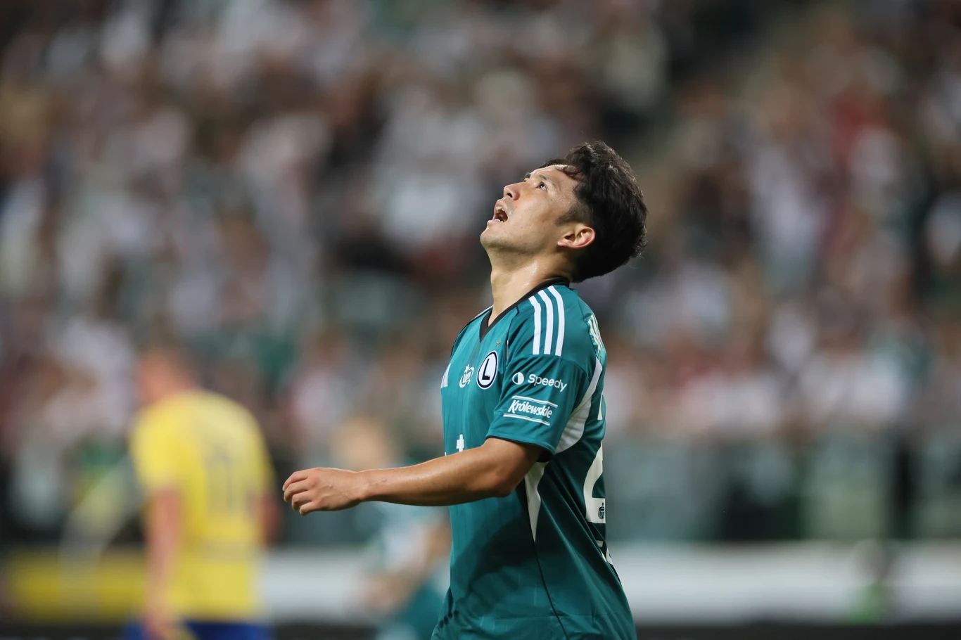 Legia Warszawa przegrała 1:4 z AEK Larnaka Piłkarz w zielonej koszulce patrzący w górę ze zmęczoną lub rozczarowaną miną na tle rozmytego stadionu i tłumu kibiców