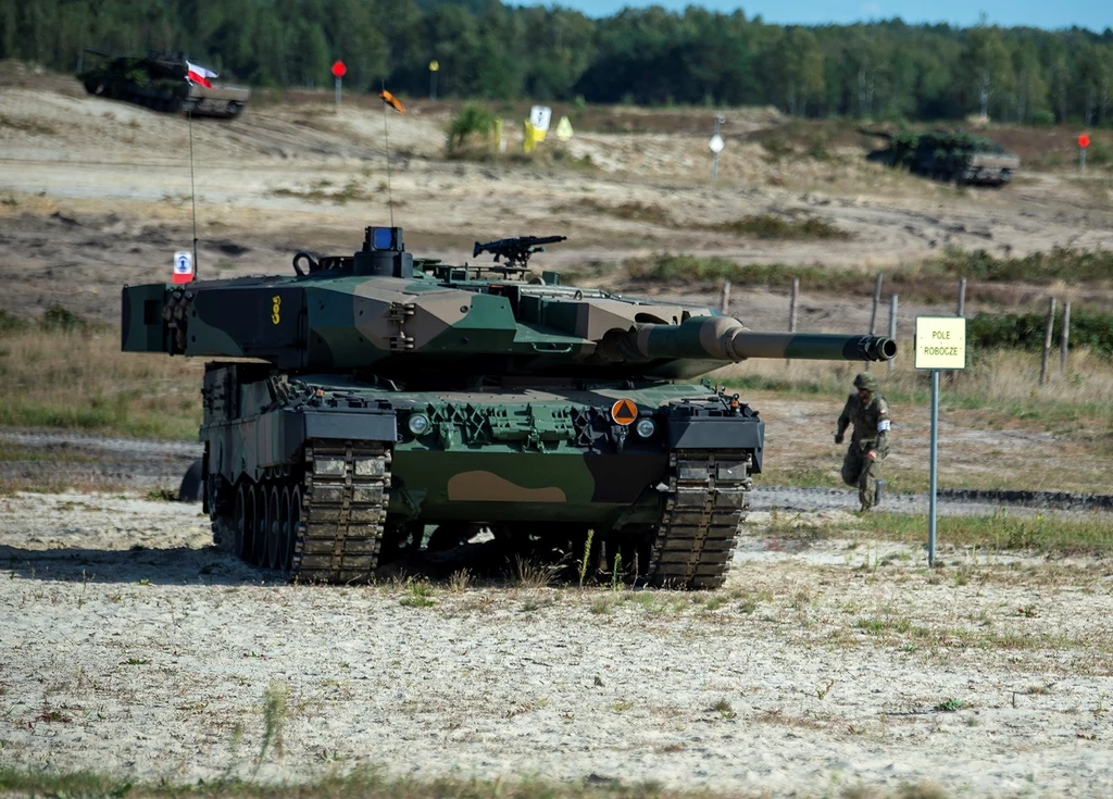 Czołg bojowy na poligonie wojskowym ustawiony naprzeciwko małego żółtego znaku, na drugim planie żołnierz przemieszczający się w stronę pojazdu, w tle widoczna zalesiona przestrzeń oraz inny pojazd wojskowy.