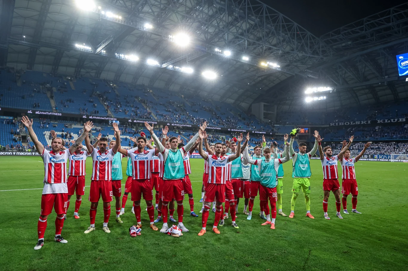 Zespół piłkarski ubrany w czerwono-białe stroje stoi na murawie stadionu, unosząc ręce w geście triumfu i radości, na trybunach widoczni kibice, a stadion jest częściowo wypełniony.