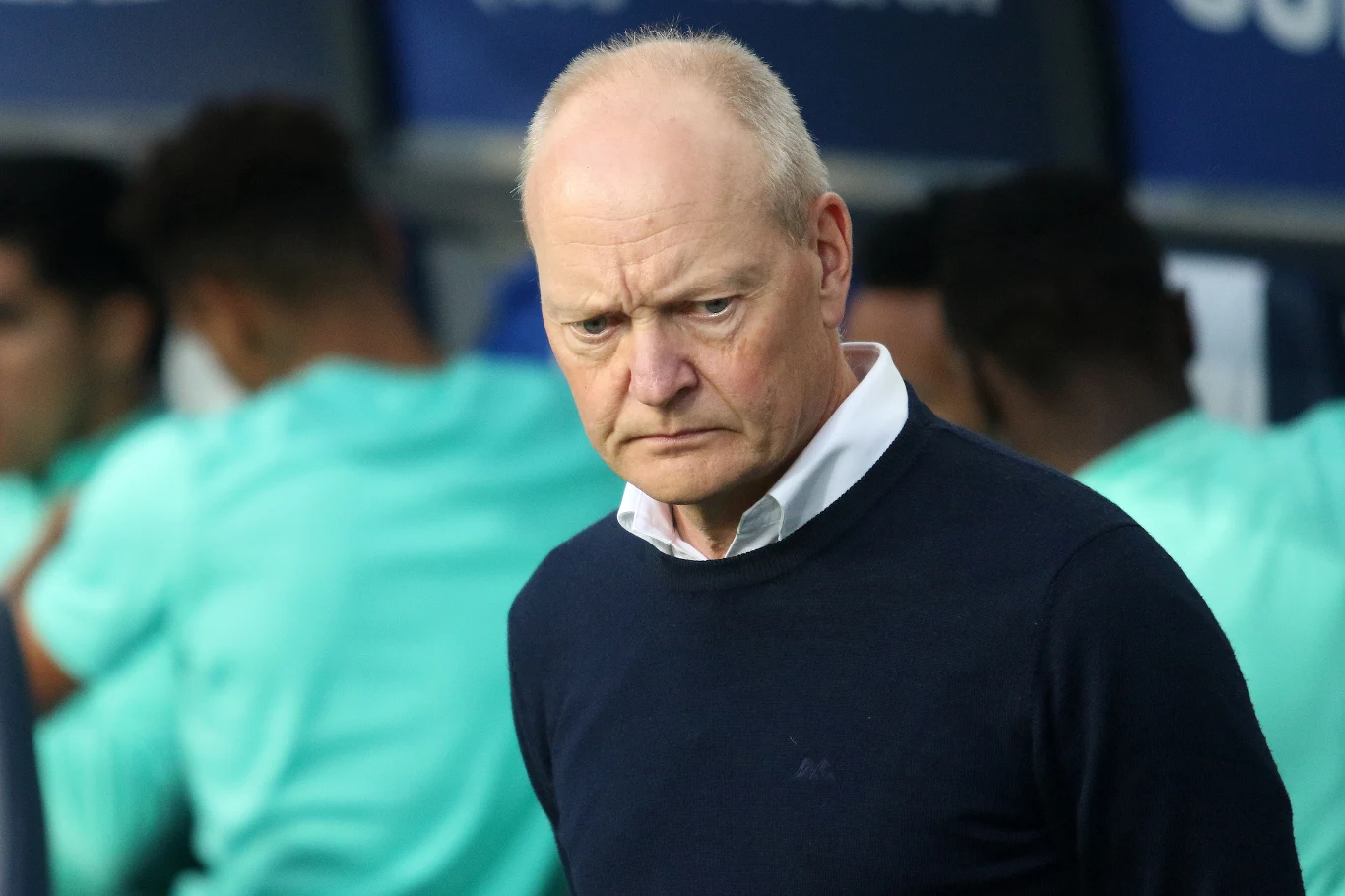 Starszy mężczyzna o poważnym wyrazie twarzy ubrany w granatowy sweter i jasnoniebieską koszulę stoi na tle rozmytych sylwetek innych osób w jasnych ubraniach, najprawdopodobniej na ławce rezerwowych podczas wydarzenia sportowego.