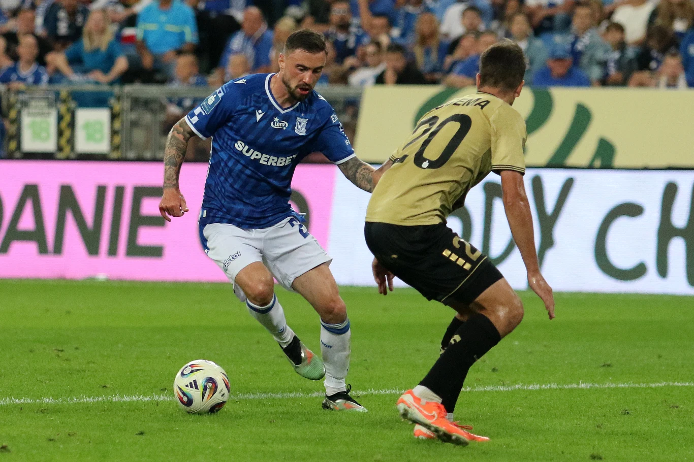 Zawodnik w niebieskiej koszulce drybluje piłkę, będąc naciskanym przez przeciwnika w złotym stroju, na tle trybun wypełnionych kibicami.