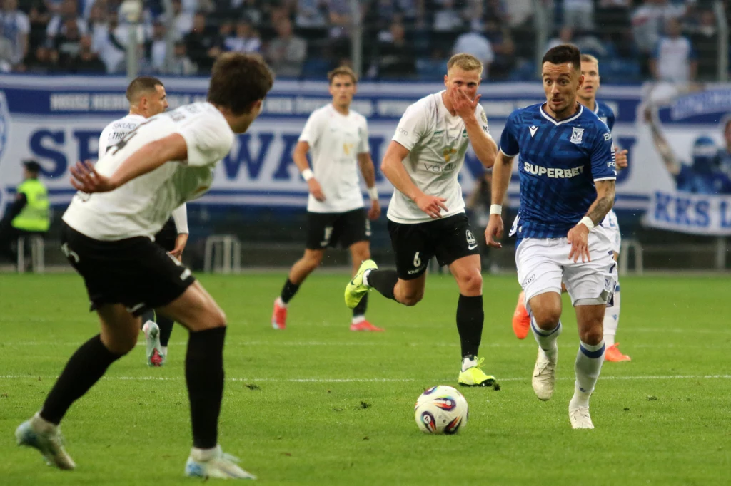 Afonso Sousa, Lech Poznań Piłkarze dwóch drużyn w trakcie rywalizacji na murawie stadionu, jeden zawodnik w niebieskiej koszulce prowadzi piłkę, pozostali podążają za akcją, widownia rozmyta w tle.
