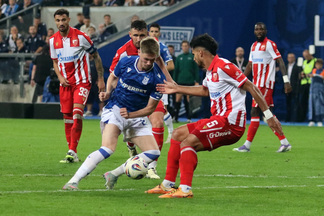 Zawodnik w niebieskiej koszulce drużyny Lecha Poznań walczy o piłkę z piłkarzem w czerwono-białym stroju, na tle stadionu wypełnionego kibicami, wokół stoją inni gracze i sędzia.