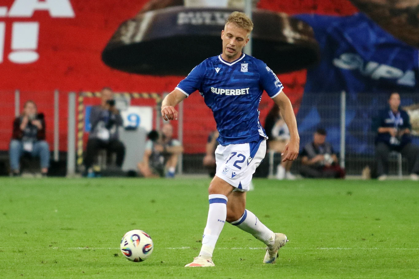 Piłkarz drużyny Lech Poznań w niebieskiej koszulce i białych spodenkach kopie piłkę na murawie stadionu podczas meczu, w tle widoczni są kibice oraz fragmenty trybun.