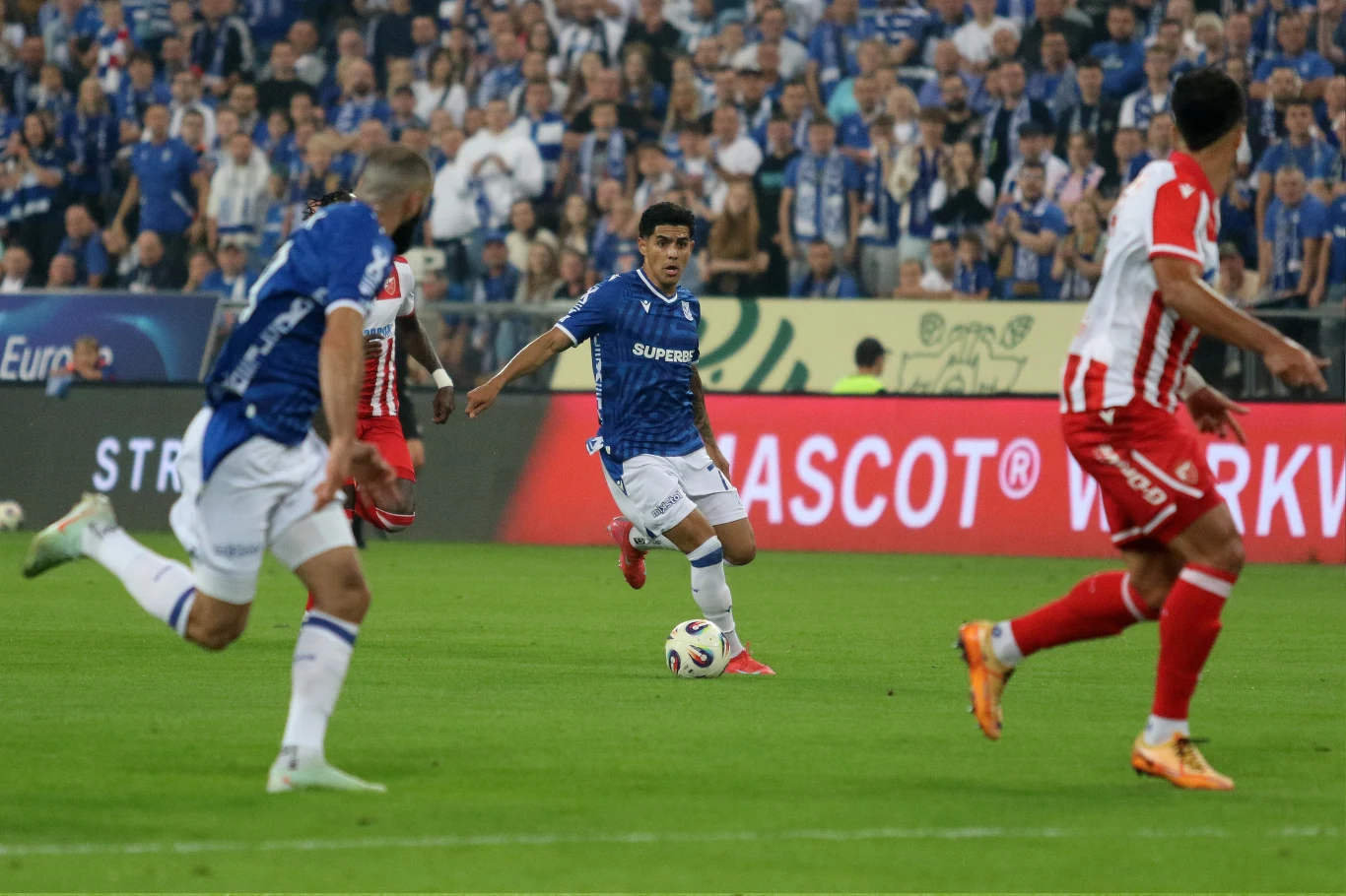 Lech Poznań przegrał z Crveną Zvezdą Piłkarze walczący o piłkę podczas dynamicznego meczu piłki nożnej, zawodnicy drużyn w niebieskich i czerwono-białych strojach, na tle wypełnionych kibicami trybun i tablic reklamowych