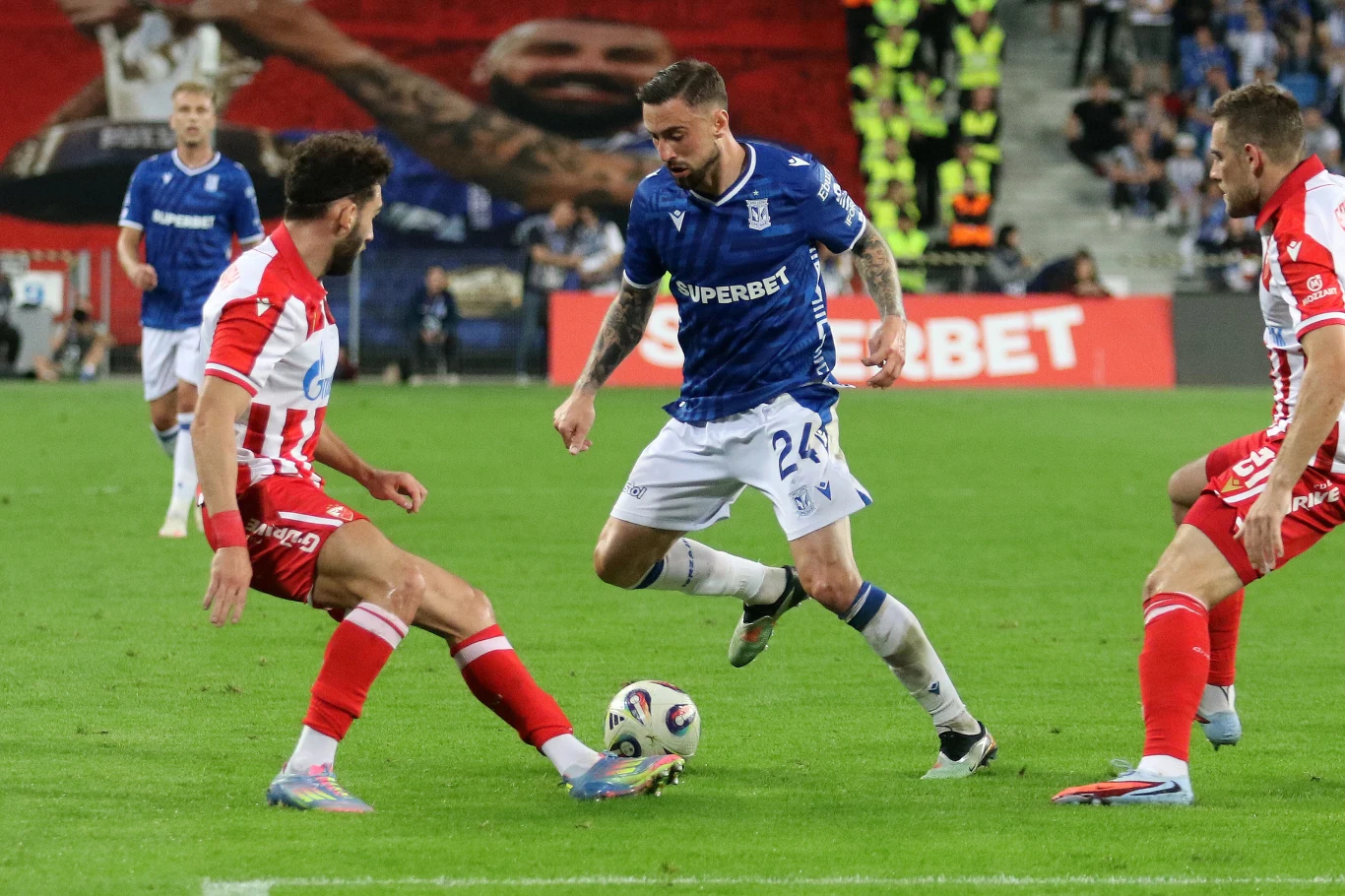 Lech Poznań nie może mówić o łatwym losie w trzeciej rudzie kwalifikacji do tegorocznej Ligi Mistrzów Piłkarze w trakcie meczu piłki nożnej walczą o piłkę na boisku, zawodnik w niebieskiej koszulce próbuje przejść między dwoma zawodnikami w biało-czerwonych strojach, w tle widownia oraz osoby asekurujące wydarzenie.