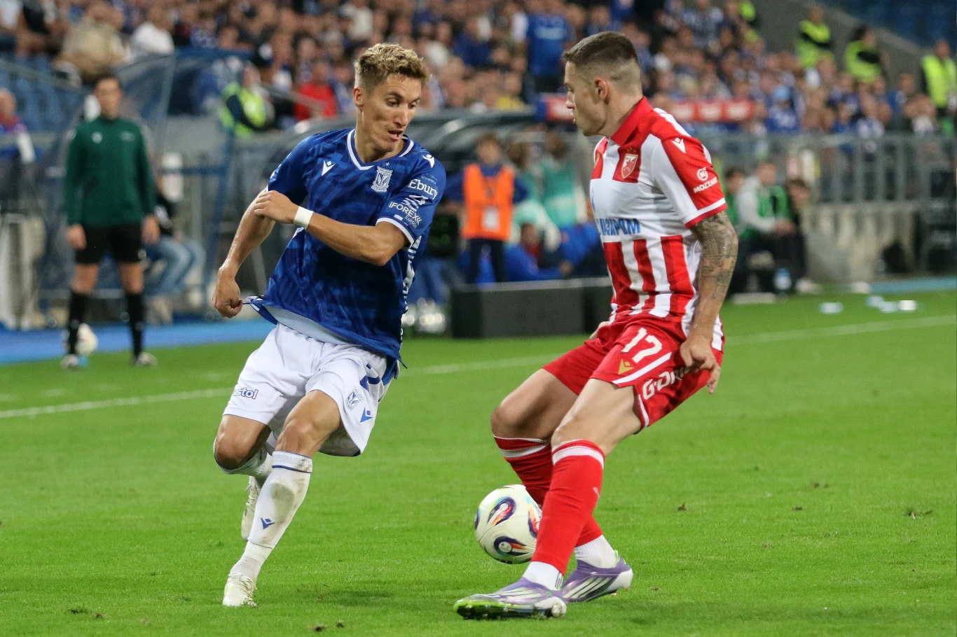 Dwóch piłkarzy rywalizuje o piłkę podczas meczu na stadionie, jeden ubrany w niebieską koszulkę i białe spodenki, drugi w czerwono-białe stroje, w tle trybuny wypełnione kibicami.