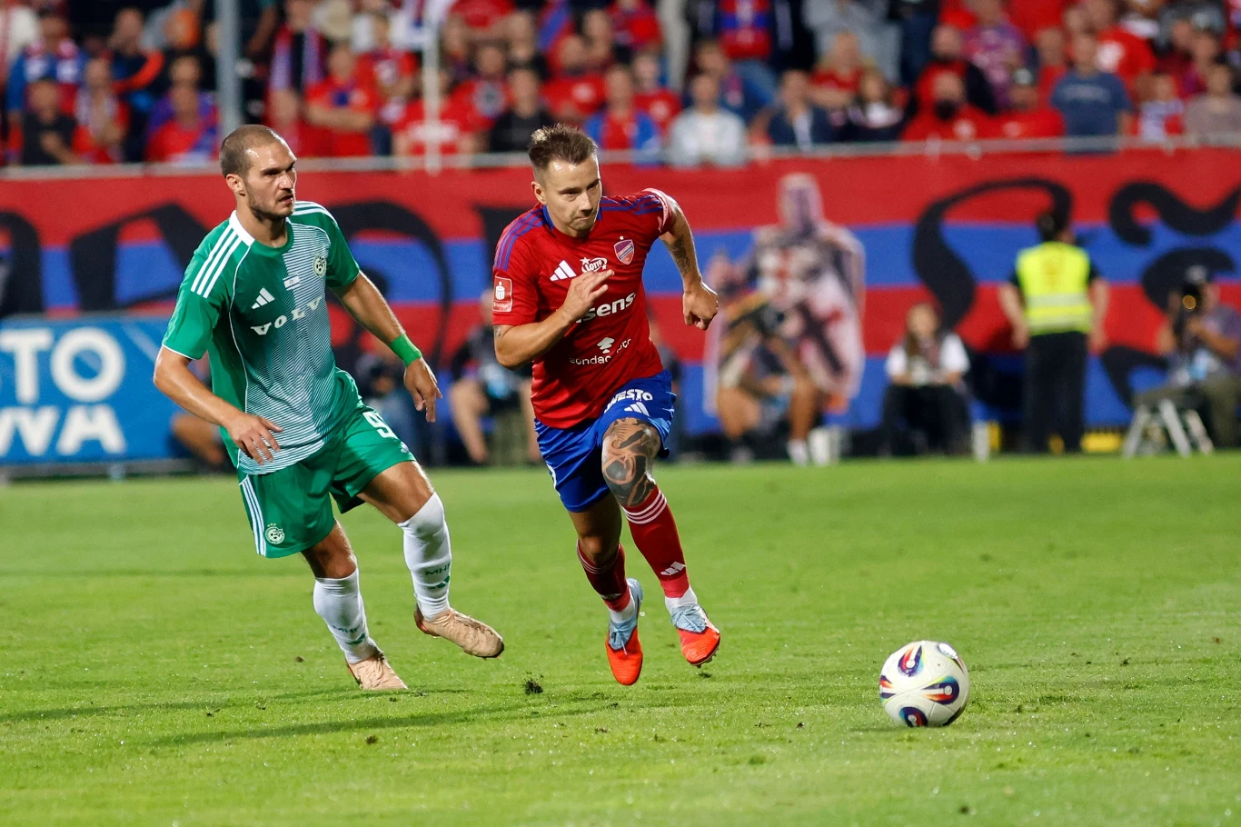 Mecz Raków Częstochowa - Maccabi Hajfa Dwóch piłkarzy rywalizuje o piłkę na boisku, jeden ubrany w zielony, drugi w czerwono-niebieski strój, w tle trybuny wypełnione kibicami oraz banerami klubowymi.