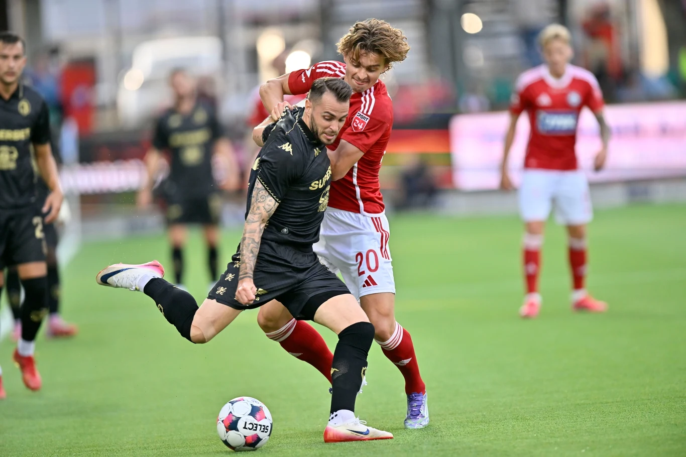 Mecz Silkeborg - Jagiellonia Białystok Dwóch piłkarzy rywalizujących o piłkę podczas meczu na murawie, zawodnik w czarnym stroju stara się utrzymać kontrolę nad piłką, gdy drugi w czerwono-białym próbuje go powstrzymać, w tle widoczni inni zawodnicy.