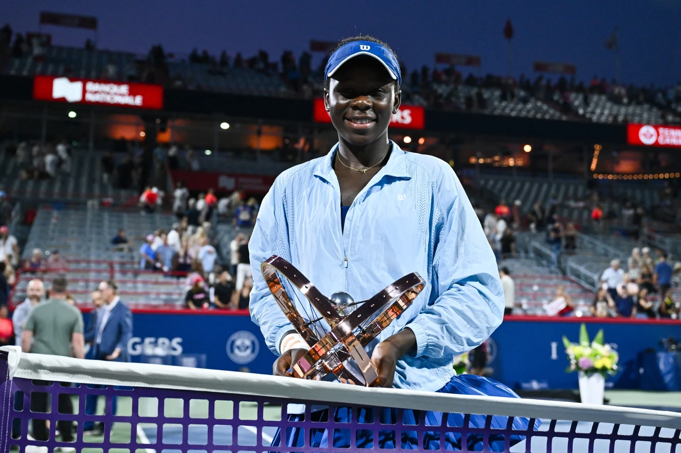 Młoda kobieta ubrana w sportową niebieską kurtkę i czapkę z daszkiem stoi przy siatce kortu tenisowego, trzymając w rękach trofeum turniejowe, w tle widoczni kibice na trybunach.