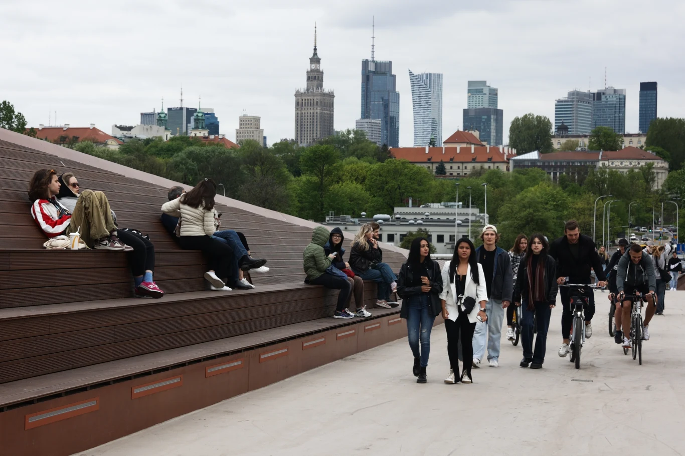 Mieszkańcy Polski stali się celem propagandy (zdj. ilustracyjne) Grupa ludzi spacerująca i siedząca na nowoczesnej, miejskiej konstrukcji z widoczną panoramą Warszawy w tle, z charakterystycznymi wieżowcami oraz Pałacem Kultury i Nauki, otoczenie zieleni i zabudowa miejską.