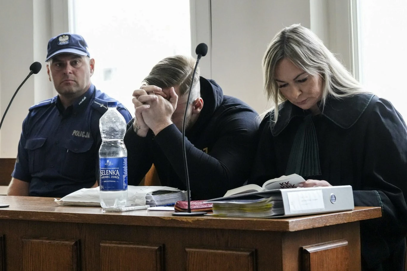 Oskarżony o spowodowanie tragicznego wypadku na Trasie Łazienkowskiej Łukasz Żak podczas procesu trzymał twarz w dłoniach Mężczyzna siedzi przy stole sądowym z głową spuszczoną i rękoma zakrywającymi twarz, obok niego kobieta czyta dokumenty, a po drugiej stronie siedzi policjant w mundurze, na stole leżą akta i butelka wody.