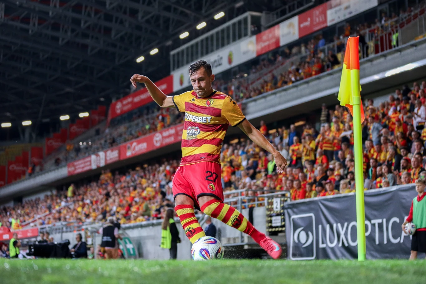 Piłkarz w żółto-czerwonym stroju oddaje rzut rożny na stadionie wypełnionym kibicami, widoczne są linie boczne boiska, chorągiewka narożna i trybuny z publicznością.